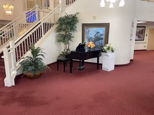 Interior lobby area with a black grand piano, floral arrangements and potted plants beneath a staircase on red carpet.