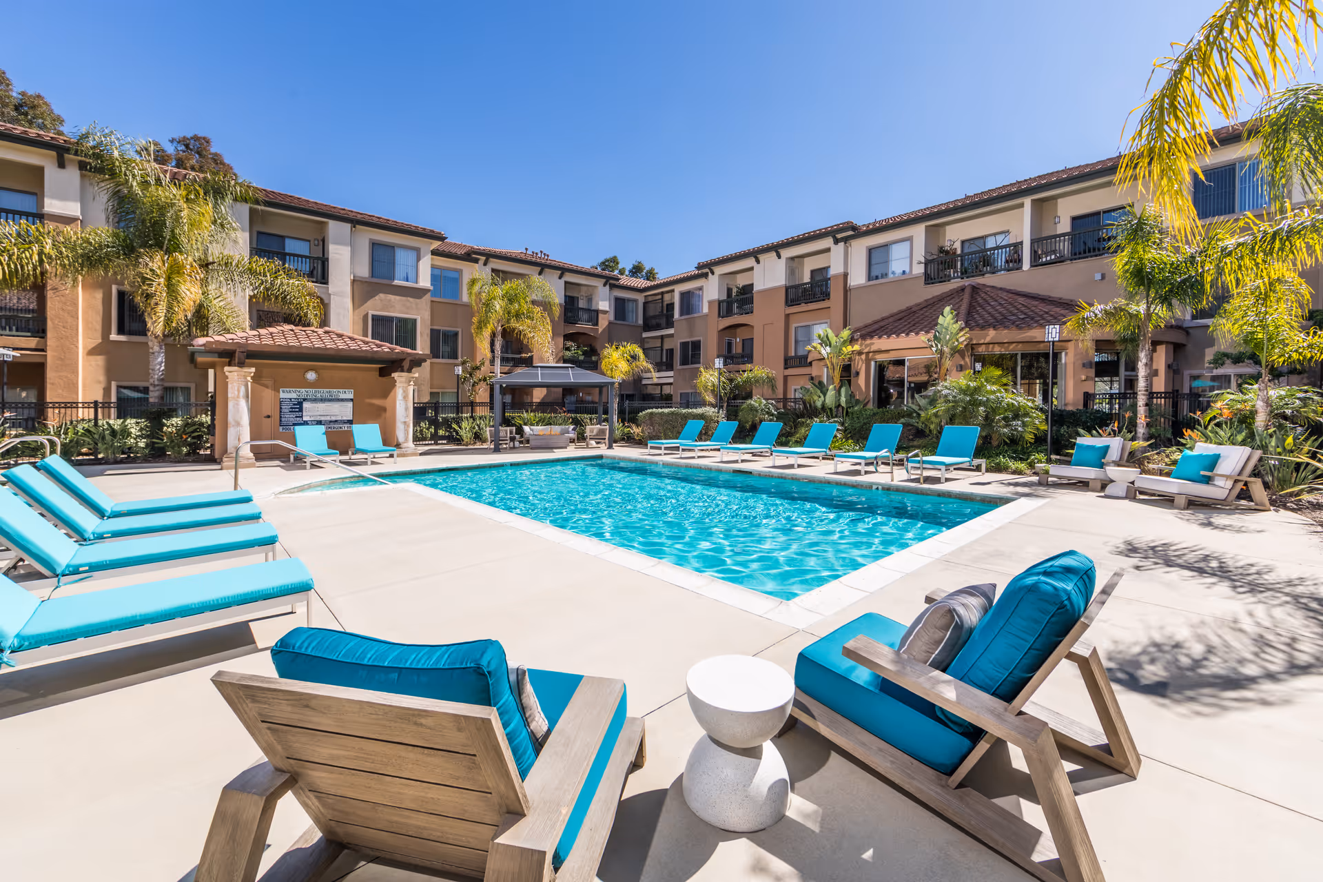 Outdoor swimming pool area at a senior living facility with turquoise cushioned lounge chairs and wooden chairs surrounding the pool. The pool is set in a courtyard with a three-story building in the background, palm trees, and clear blue sky.