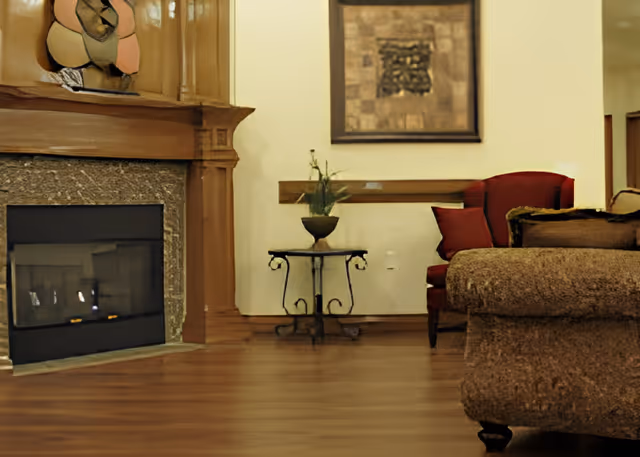 Cozy living room area with a stone and wood fireplace on the left, a small decorative table with a plant in the center, and a red armchair with cushions next to a brown upholstered sofa on the right. A framed abstract artwork hangs on the wall above the table.