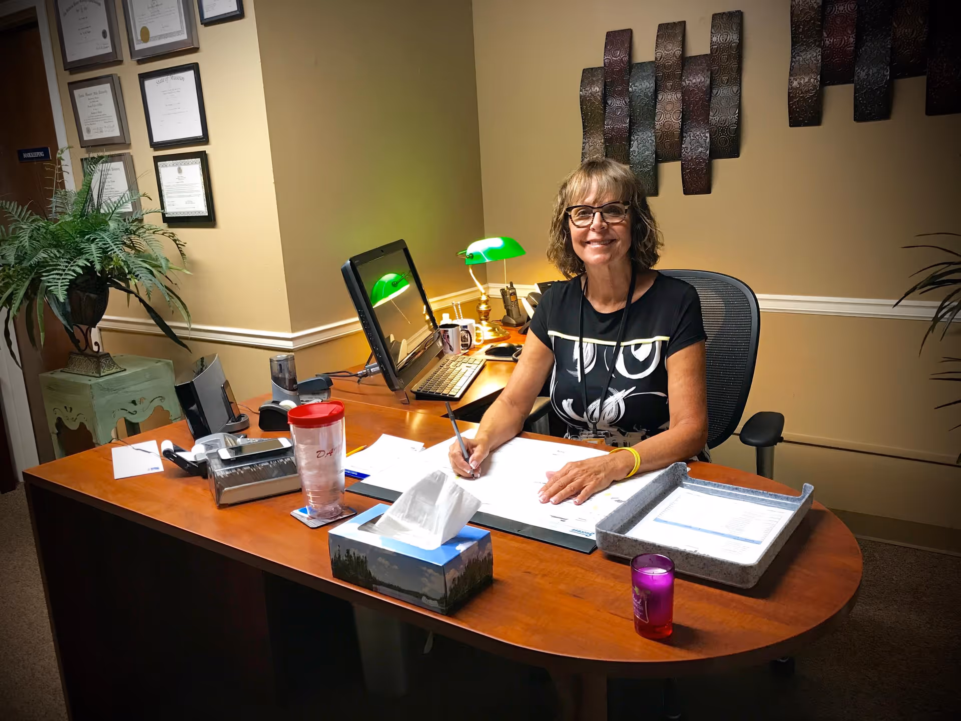 A woman with glasses sitting at a wooden desk in an office, smiling while writing on a paper. The desk has a computer monitor, a green desk lamp, a tissue box, a red cup, and some office supplies. Certificates are hanging on the wall behind her, and there is a plant on a small table to the left.