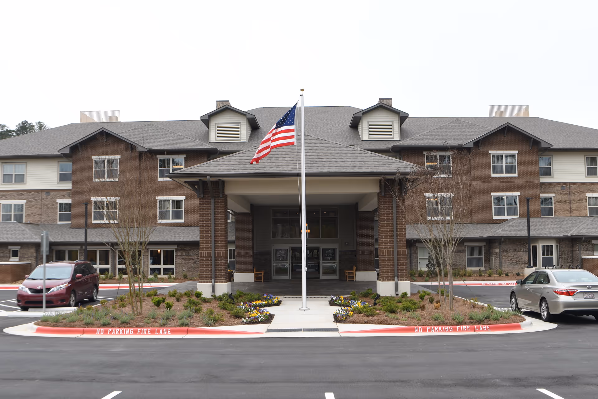 Front exterior view of Brier Pointe Retirement Community building with a covered entrance, an American flag on a flagpole in the center, landscaped flower beds, and parked cars on either side.