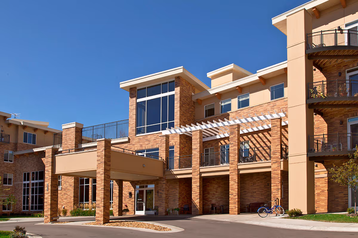 Exterior view of Carondelet Village senior living facility showing a multi-story brick building with large windows, balconies, and a covered entrance under a clear blue sky.