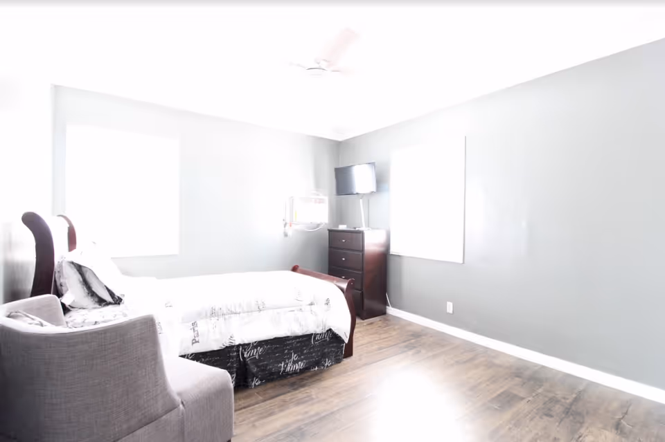 Bright bedroom with a single bed, an armchair, a chest of drawers with a small TV, and gray walls.