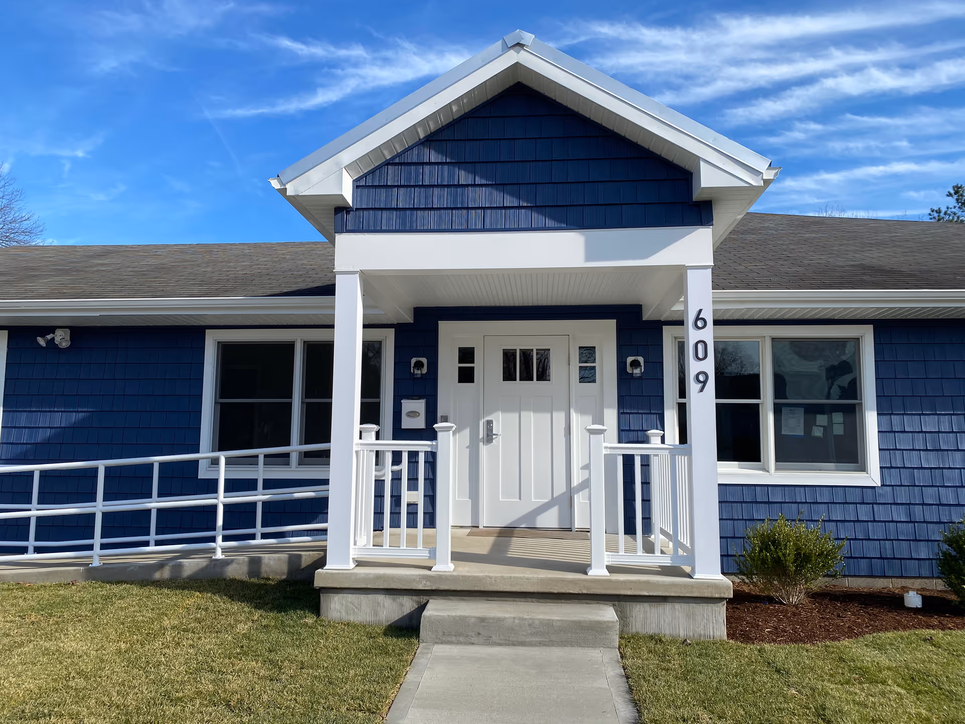 Front exterior view of a single-story building with blue siding and white trim. The entrance has a small covered porch with white railings and a ramp on the left side. The building number 609 is displayed vertically on a white column next to the door. The sky is clear and blue.