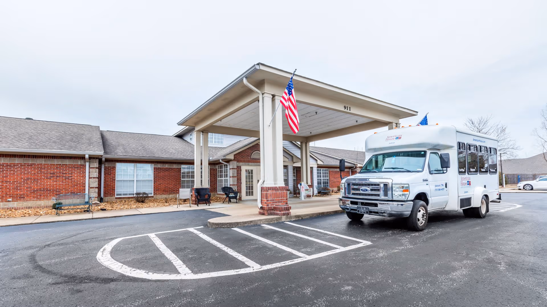 Exterior view of Wellington Manor Jackson showing the entrance with a covered drop-off area, an American flag hanging from the structure, and a white shuttle bus parked in front. The building is made of red brick with white trim and has several windows.