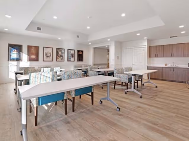 A bright, spacious room with multiple tables and cushioned chairs arranged neatly, featuring light wood flooring, white walls, framed artwork on the wall, and a kitchenette area with wooden cabinets and a sink in the background.