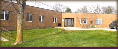 Single-story brick facility with glass entry, surrounding lawn, tree, and parked car.