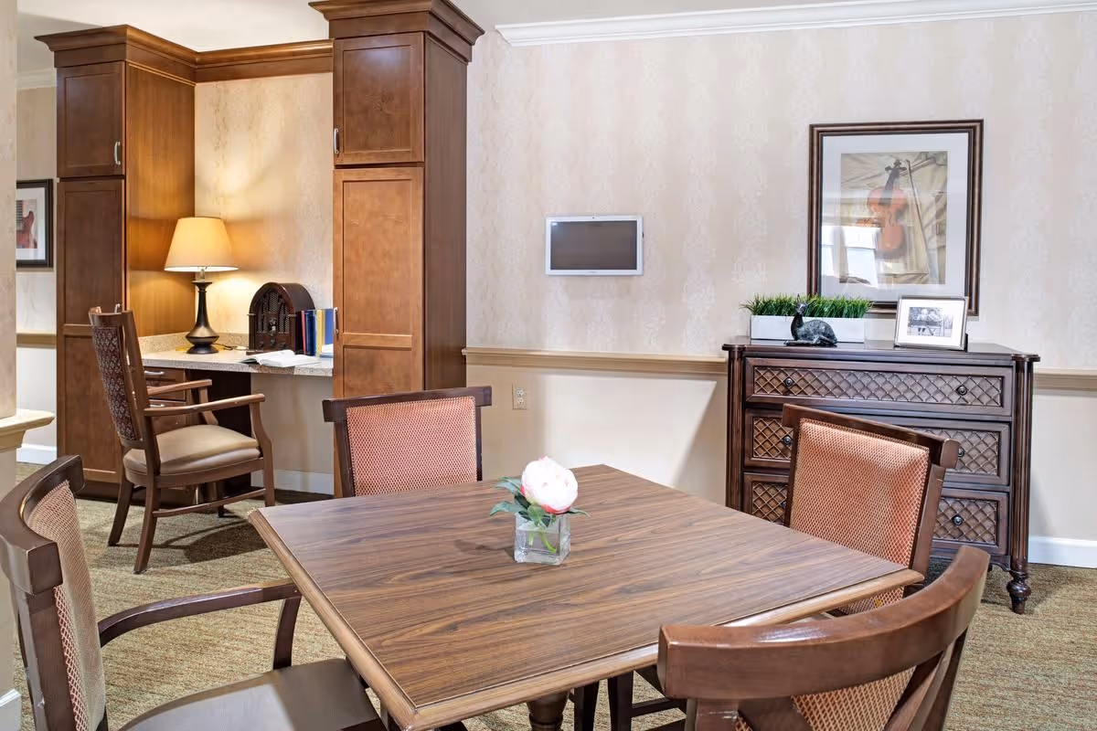 Wooden dining table with chairs in a furnished common room featuring a sideboard, desk alcove with a lamp, and framed artwork.