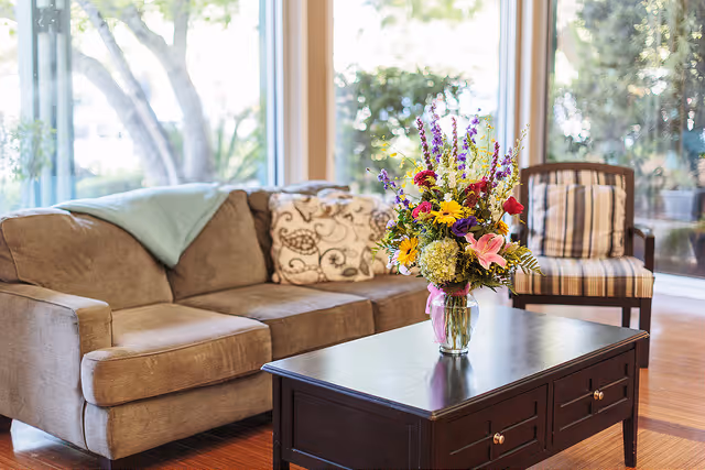 A cozy living room area with a brown sofa adorned with a patterned pillow and a light blue throw blanket. In front of the sofa is a dark wooden coffee table with a colorful bouquet of flowers in a glass vase. A striped armchair is positioned near large windows that let in natural light and offer a view of greenery outside.