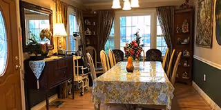 A dining room with a rectangular table covered with a floral tablecloth and a vase of flowers in the center. The room has wooden chairs around the table, a sideboard with a lamp and decorative items, and built-in shelves with various ornaments. French doors with glass panes lead outside, and the room is warmly lit with ceiling lights.