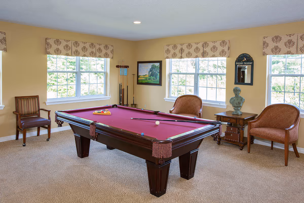 A well-lit game room with a burgundy pool table in the center, two pool cues resting on it, and a rack of billiard balls arranged for a game. The room has beige walls, three large windows with patterned valances, two upholstered chairs, a wooden chair, a small side table with a decorative bust, and a wall-mounted mirror with a sign above it that reads 'Billiard Academy'.