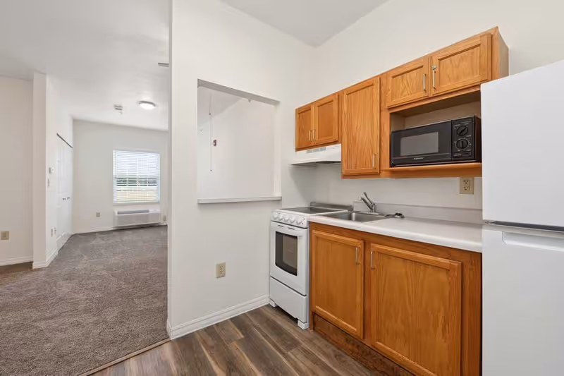 A small kitchen area with wooden cabinets, a white stove, a microwave, a sink, and a refrigerator. The kitchen opens into a carpeted living space with a window and an air conditioning unit below it.