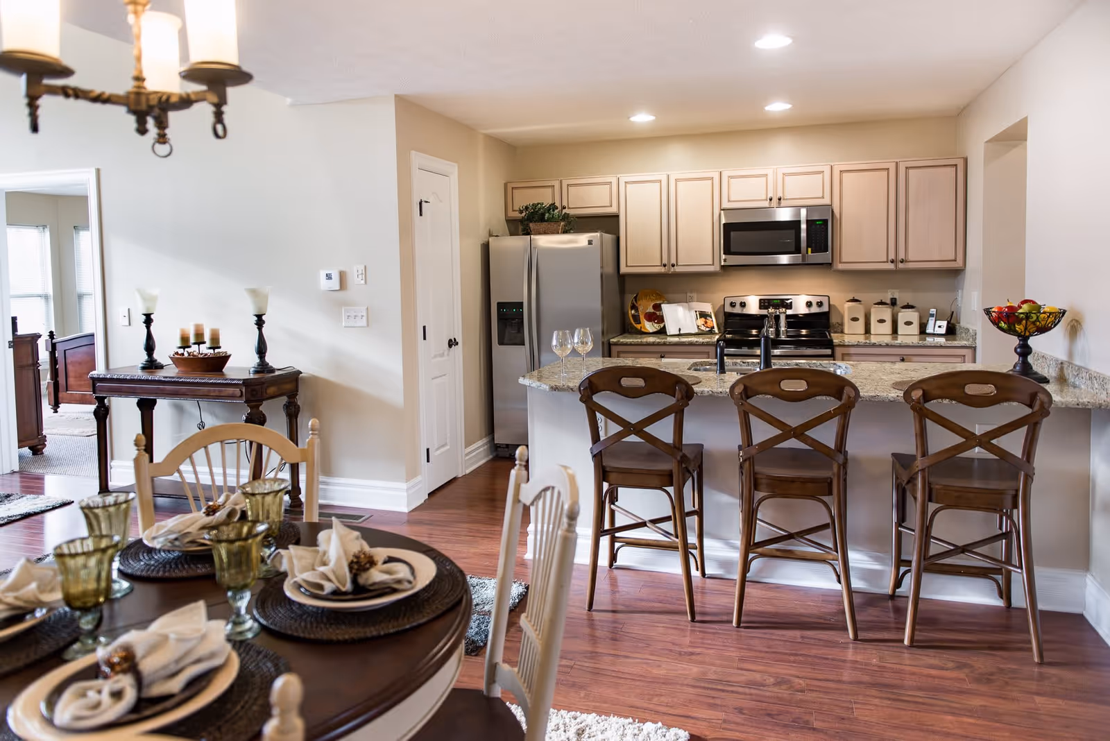 A bright and cozy kitchen and dining area with wooden flooring. The kitchen features light-colored cabinets, a stainless steel refrigerator, microwave, and stove. There is a granite countertop island with three wooden bar stools and two wine glasses on the counter. The dining area has a round wooden table set with plates, napkins, and green-tinted glasses. A decorative table with candles and lamps is visible against the wall, and a bedroom can be seen through an open doorway.