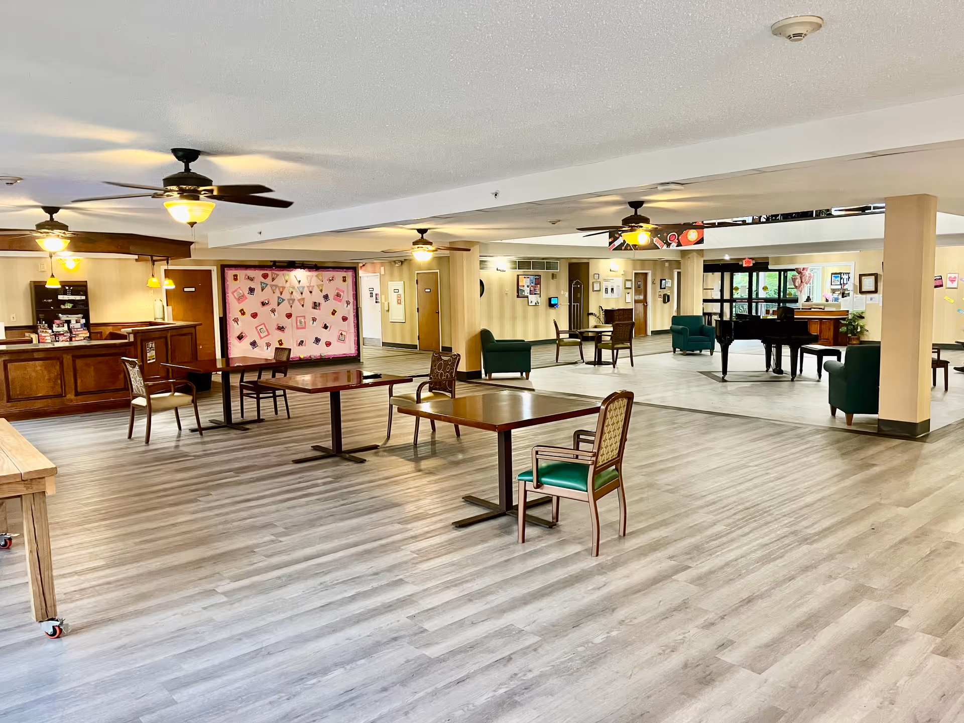 Spacious common area in a senior living facility with wooden tables and chairs, ceiling fans with lights, a grand piano, and green armchairs. The room has light-colored wood flooring and beige walls with various decorations and a bulletin board.