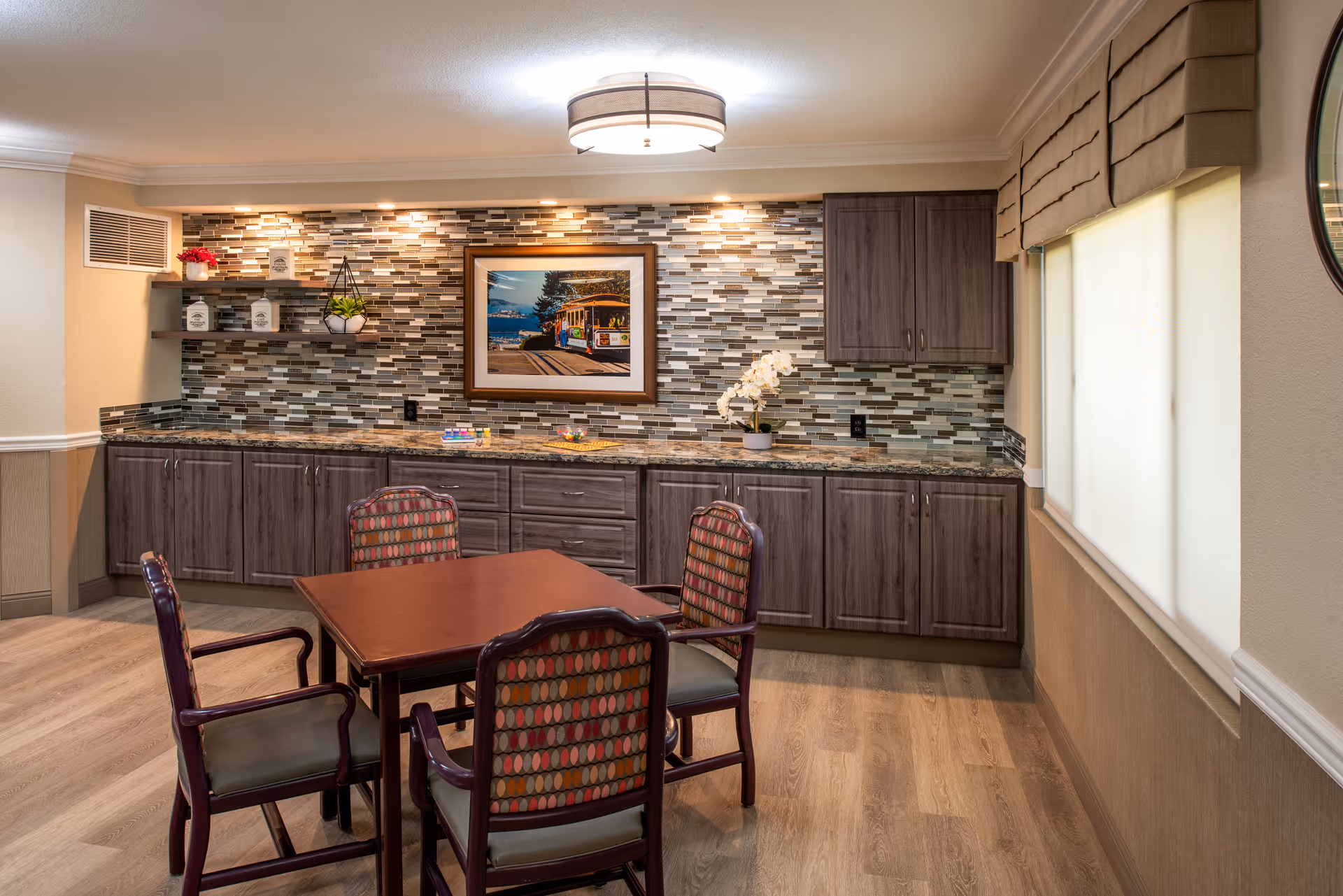 A cozy interior room with a wooden table and four patterned chairs in the center. The back wall features a mosaic tile backsplash with dark wood cabinets below and above. A framed picture of a streetcar and a potted plant are displayed on the countertop. The room has wood flooring, a window with beige Roman shades on the right, and a modern ceiling light fixture.