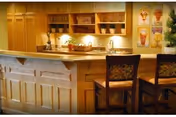 A warm, inviting kitchen area with wooden cabinetry and a countertop bar with two chairs featuring floral upholstery. The countertop has a basket and some decorative items, and the background includes shelves with baskets and framed artwork on the wall.