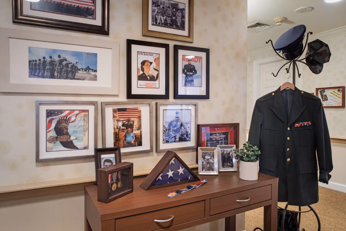 A memorial display in a senior living facility featuring a military uniform on a coat rack, a folded American flag in a wooden case, framed military medals, and several framed photographs and patriotic posters on the wall.