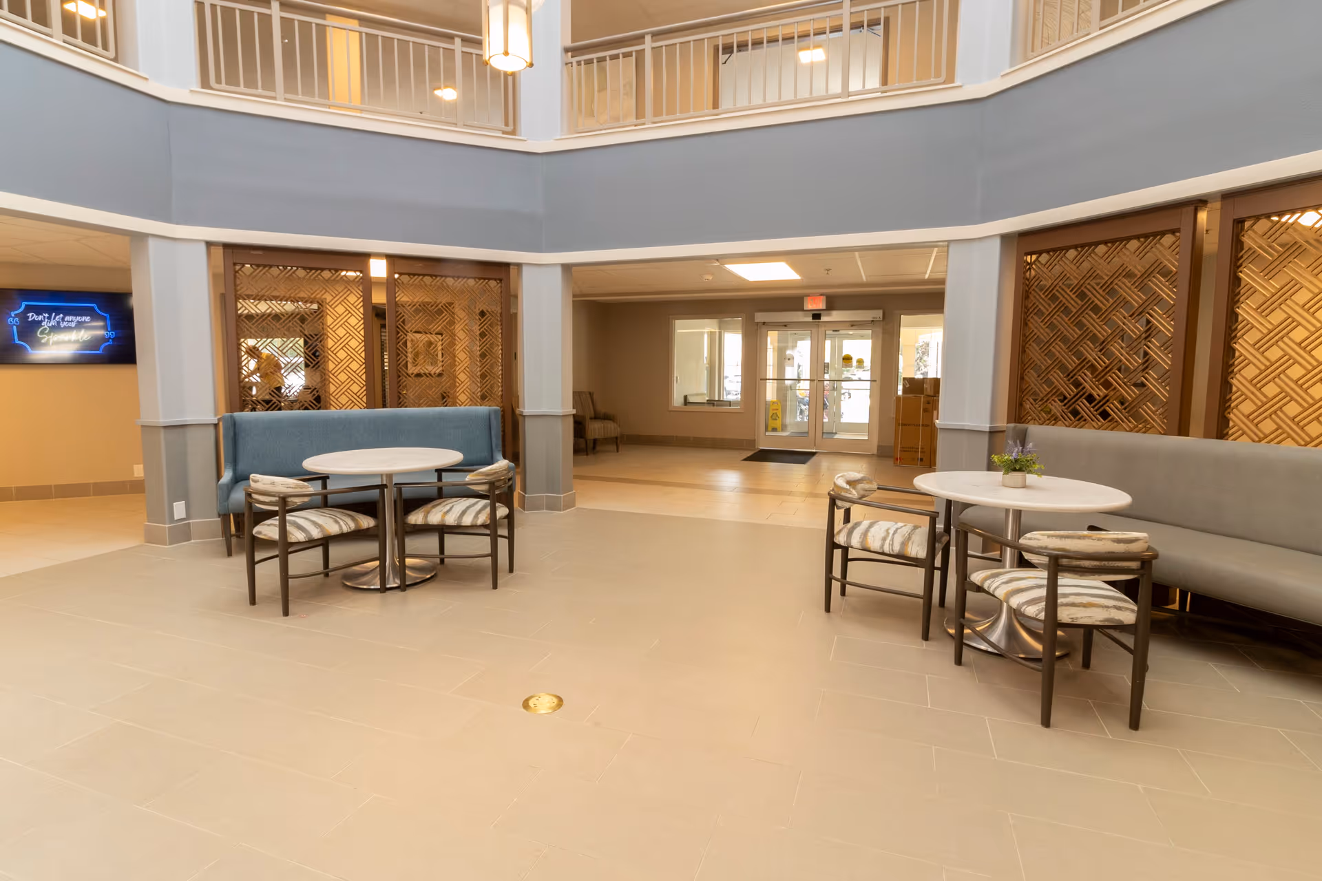 Interior view of a senior living facility common area with two round tables, each surrounded by four chairs. One table has a blue cushioned bench against the wall, and the other has a gray cushioned bench. The area has beige tiled flooring, decorative wooden panels on the walls, and a double glass door entrance in the background. The space is well-lit with ceiling lights and has a second-floor balcony overlooking the area.