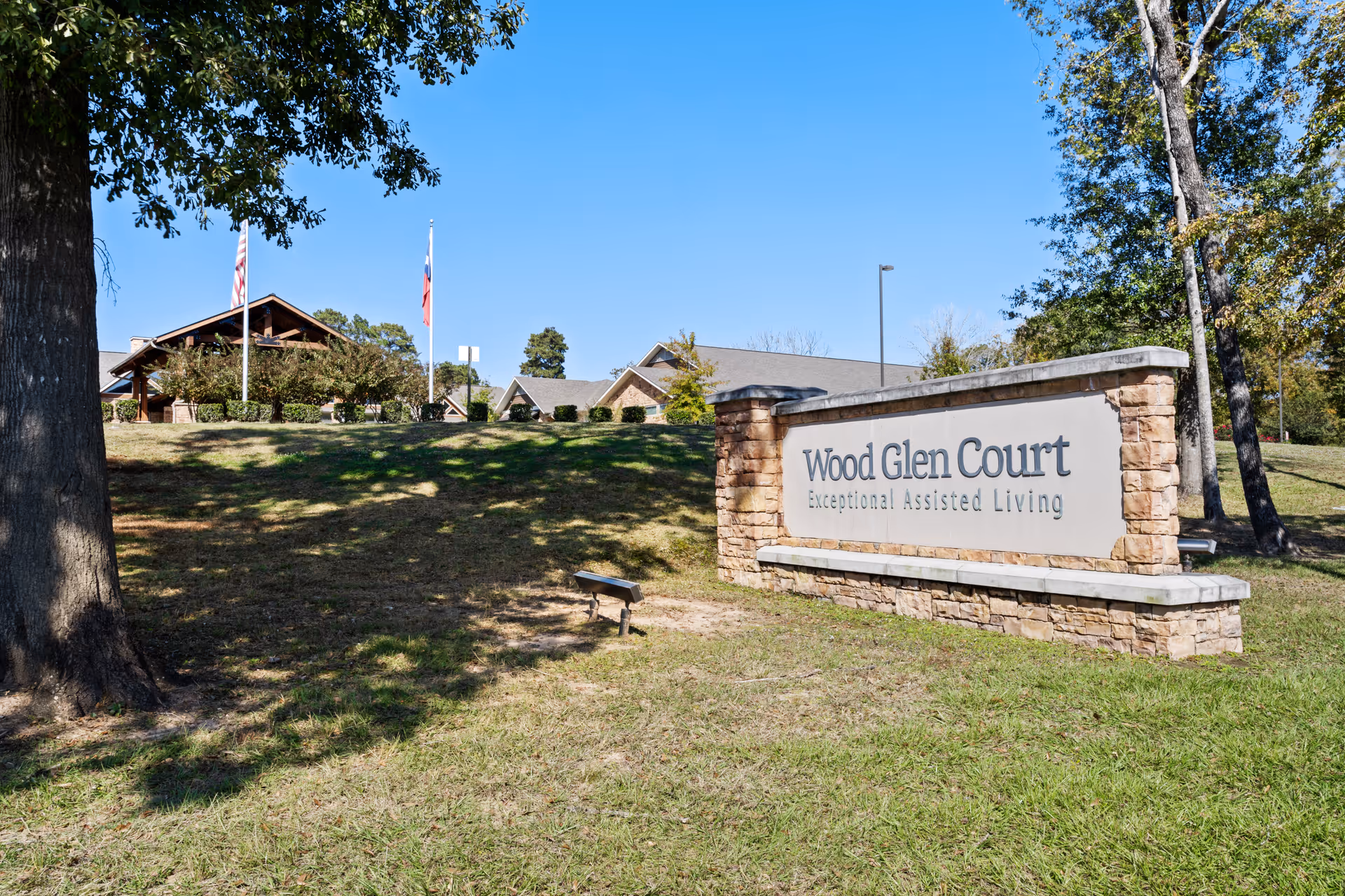 Outdoor view of Wood Glen Court assisted living facility sign made of stone and concrete with the building and flagpoles in the background under a clear blue sky.