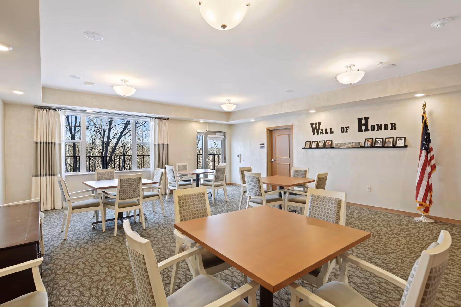 A bright and spacious room with multiple square wooden tables and cushioned chairs arranged for seating. Large windows with curtains allow natural light to fill the room, and an American flag stands in the corner. On one wall, there is a 'Wall of Honor' display with framed photos and plaques.
