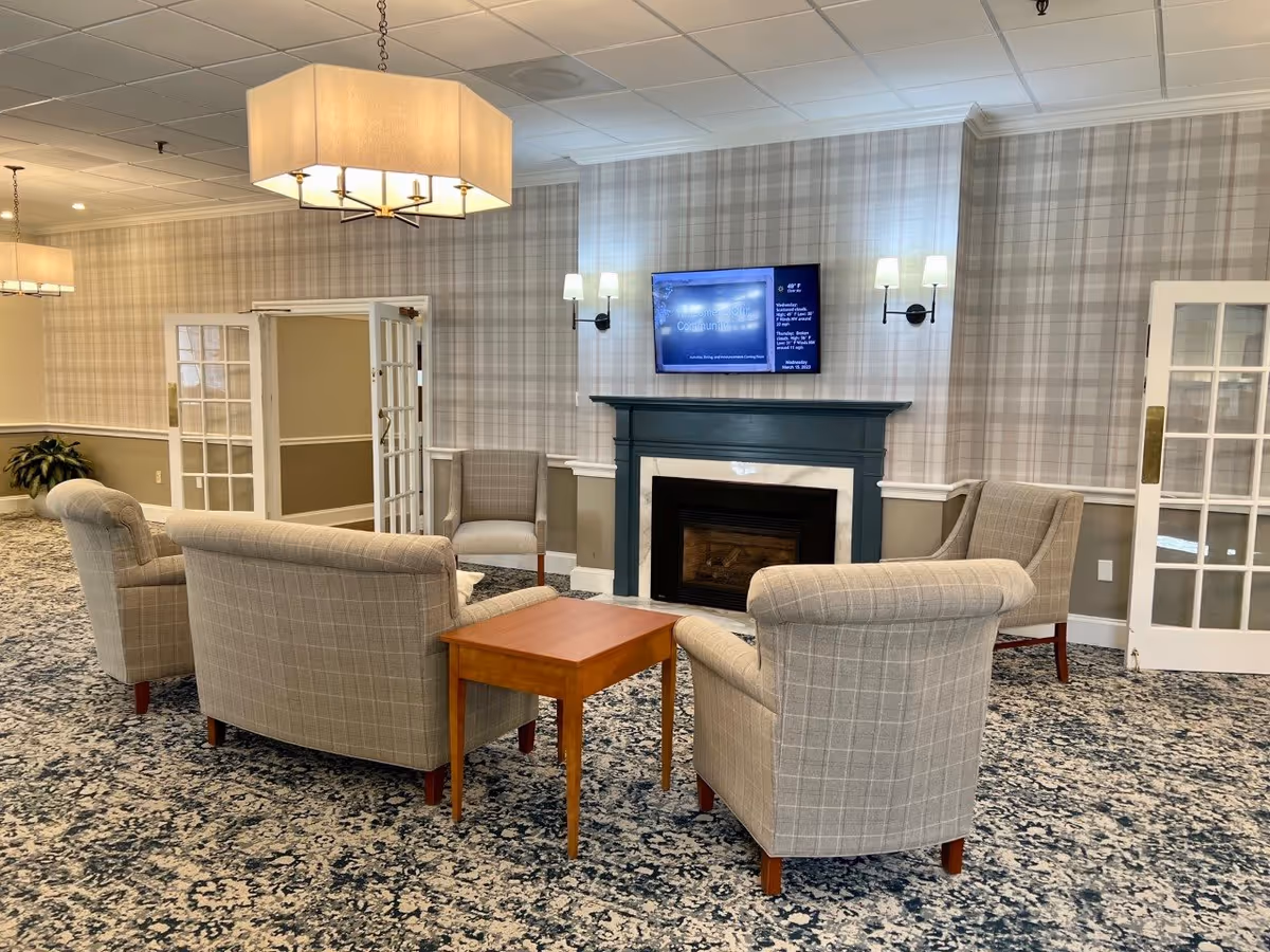 A cozy sitting area in a senior living facility with four beige plaid armchairs arranged around a wooden coffee table. The room features a fireplace with a dark green mantel, a wall-mounted TV above it, and two wall sconces on either side. The walls have a light plaid wallpaper, and there are double glass doors on both sides of the fireplace. The floor is covered with a patterned carpet, and two large ceiling lights hang overhead.