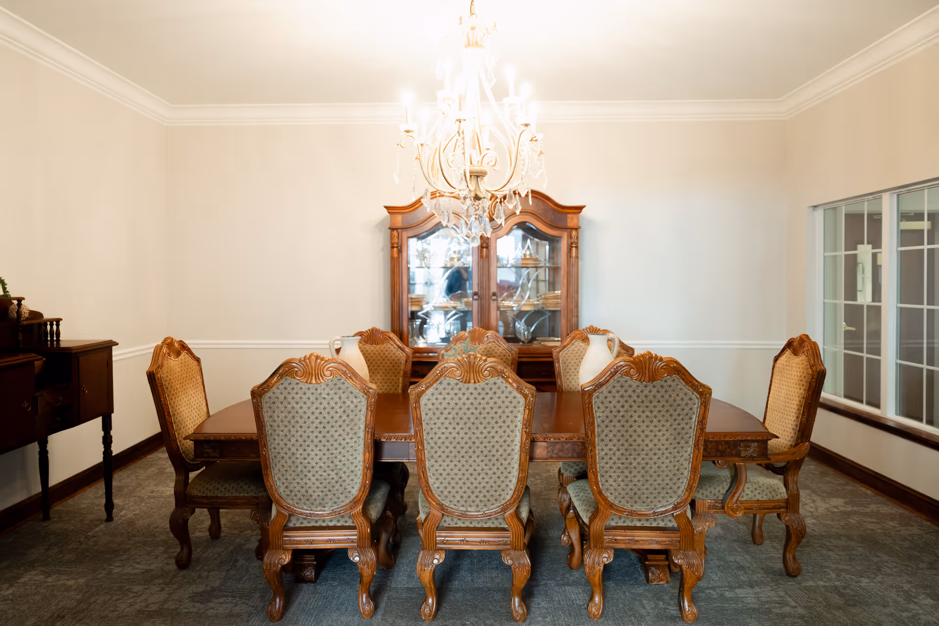 A formal dining room with a large wooden dining table surrounded by eight ornate wooden chairs with upholstered backs and seats. A crystal chandelier hangs above the table. Against the back wall is a wooden china cabinet displaying dishes and glassware. There is a window with white framing on the right side and a wooden sideboard on the left.