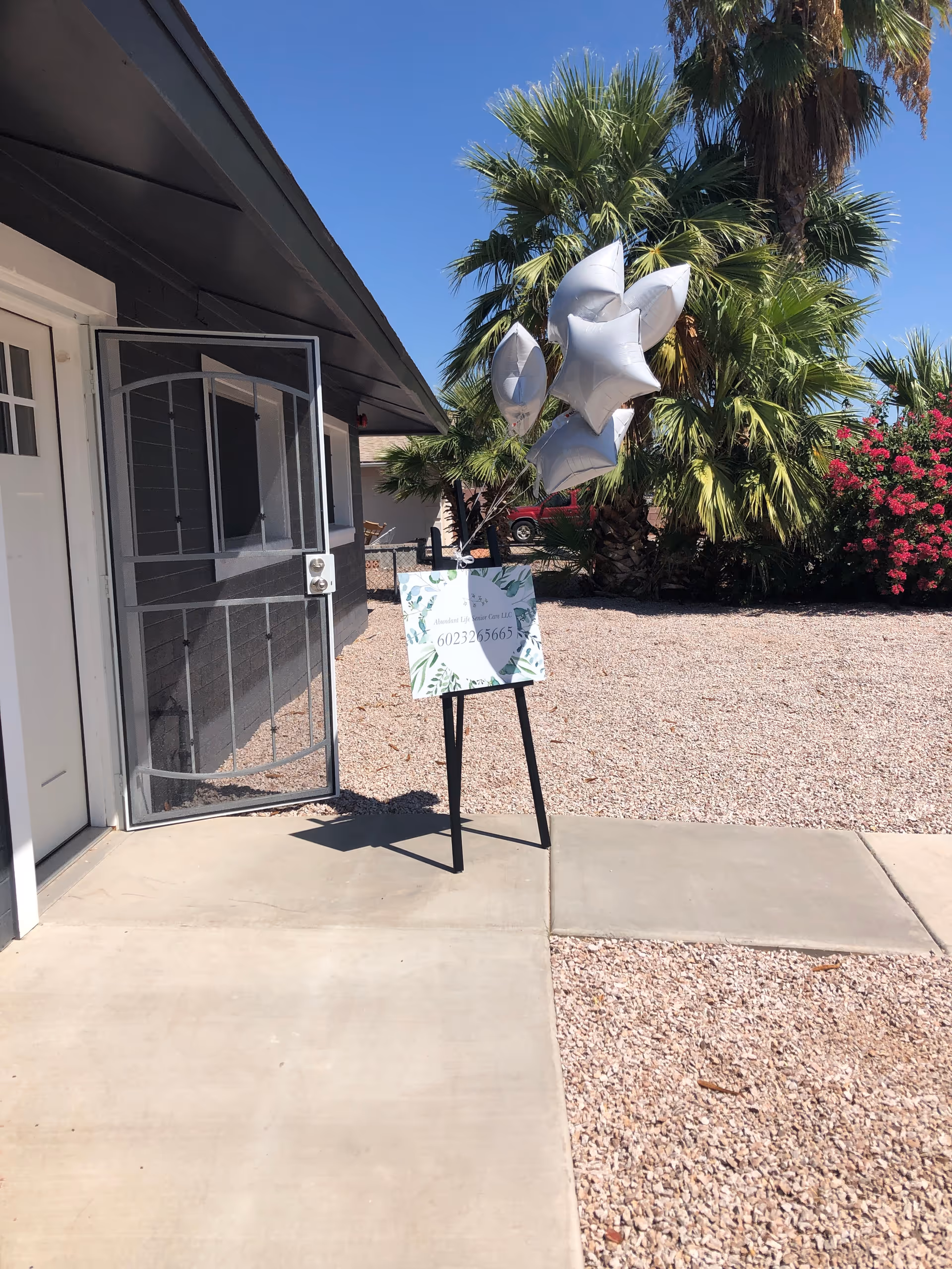 Outdoor view of the entrance to a building with a metal screen door open. A sign on a black easel with silver star-shaped balloons attached is placed on the concrete walkway. The ground is covered with gravel, and there are palm trees and flowering bushes in the background under a clear blue sky.