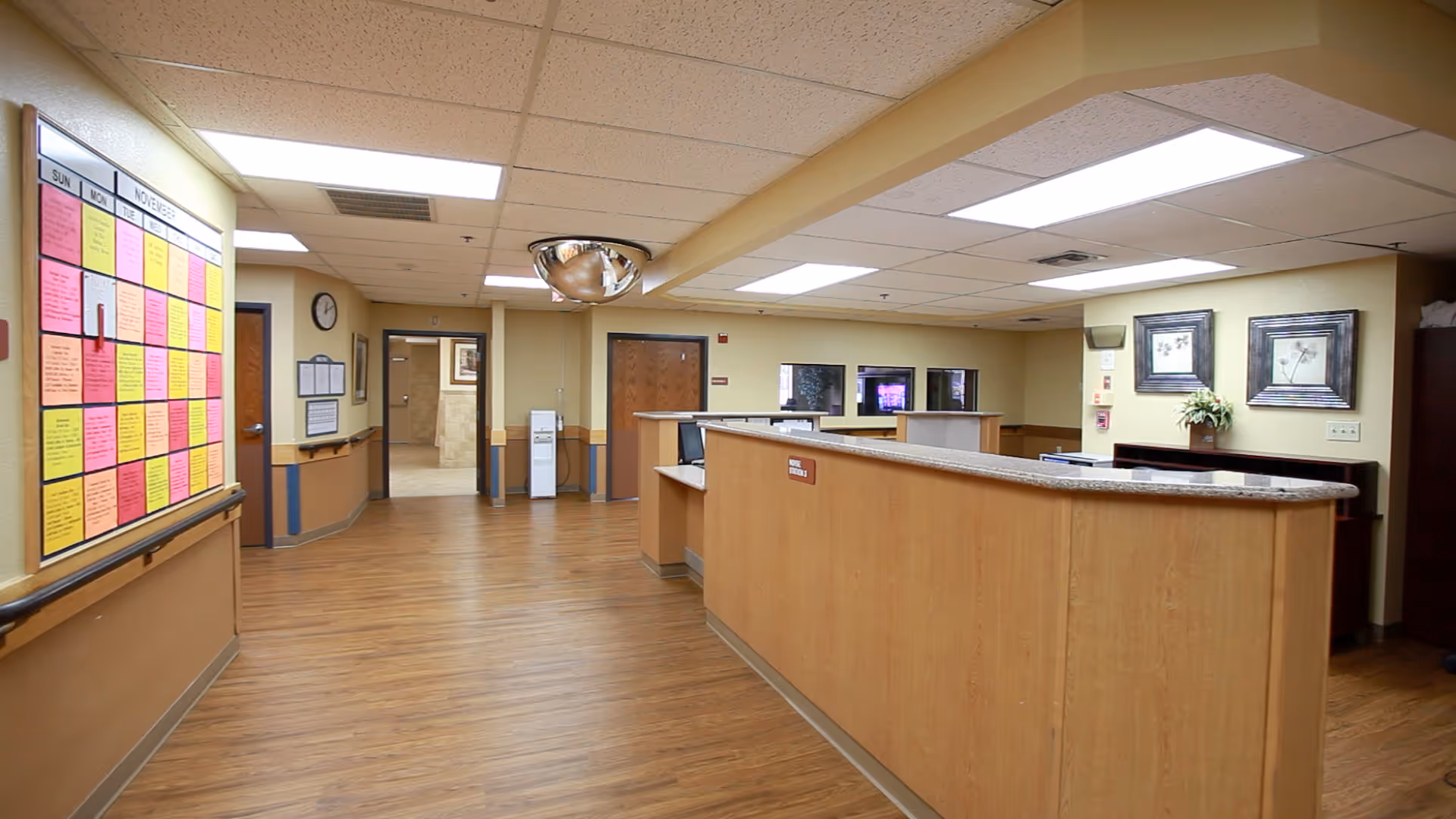 Interior view of a nursing center reception area with a wooden reception desk, a bulletin board with colorful notes on the left wall, framed pictures and a plant on the right wall, and doors leading to other rooms in the background. The floor is wooden and the ceiling has fluorescent lighting.