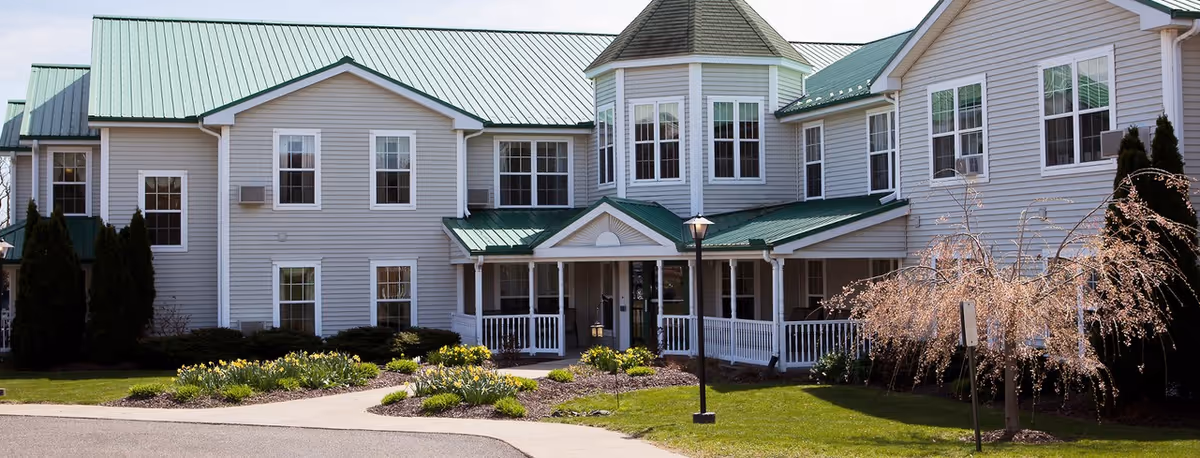 Front exterior of a two-story assisted living building with a green metal roof, white siding, covered entrance porch and landscaped grounds.