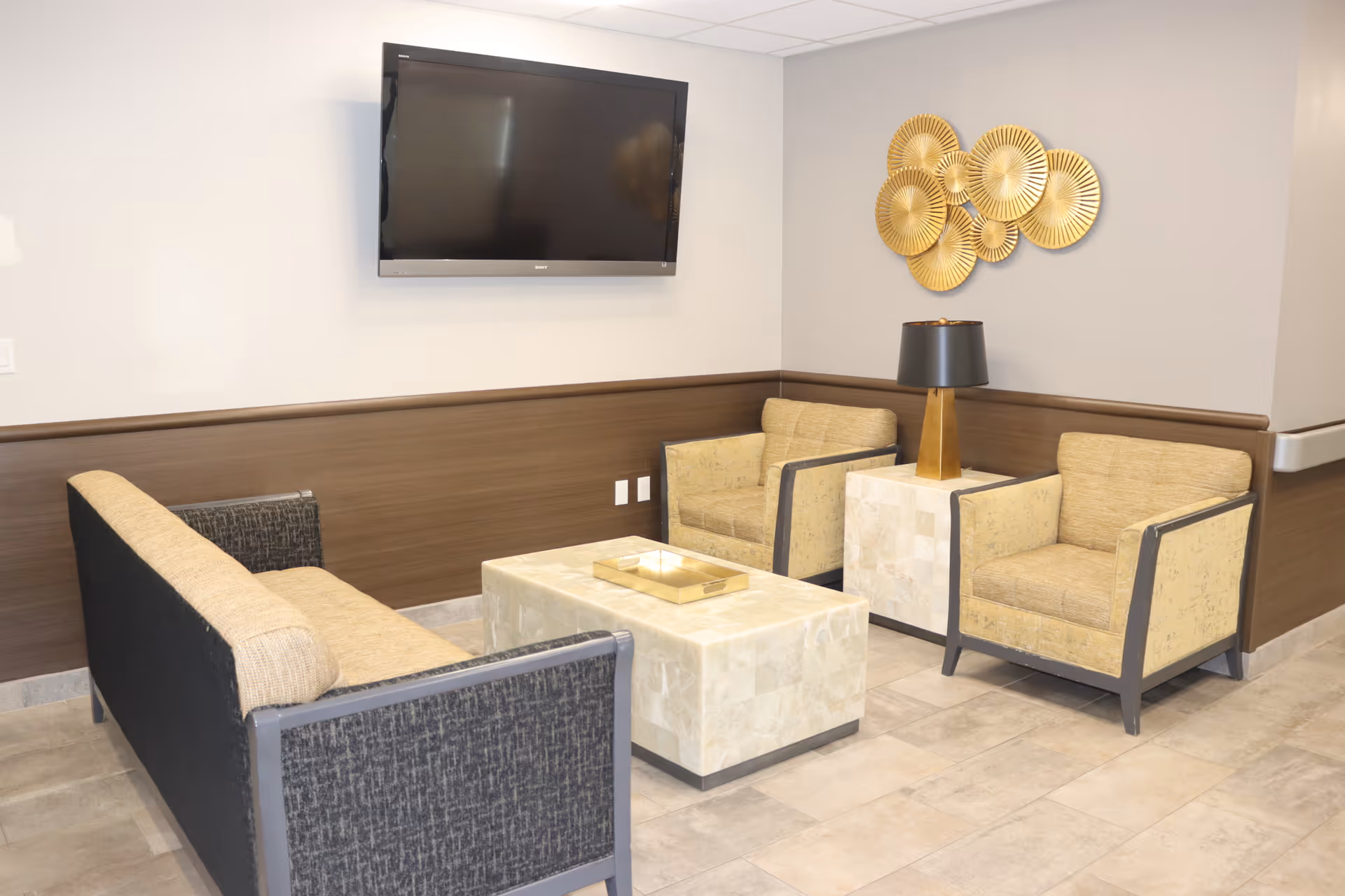 A small seating area with a beige and black sofa, two beige armchairs, a marble coffee table, and a matching side table with a black and gold lamp. A flat-screen TV is mounted on the wall above the sofa, and a decorative gold wall art piece is displayed on the adjacent wall.