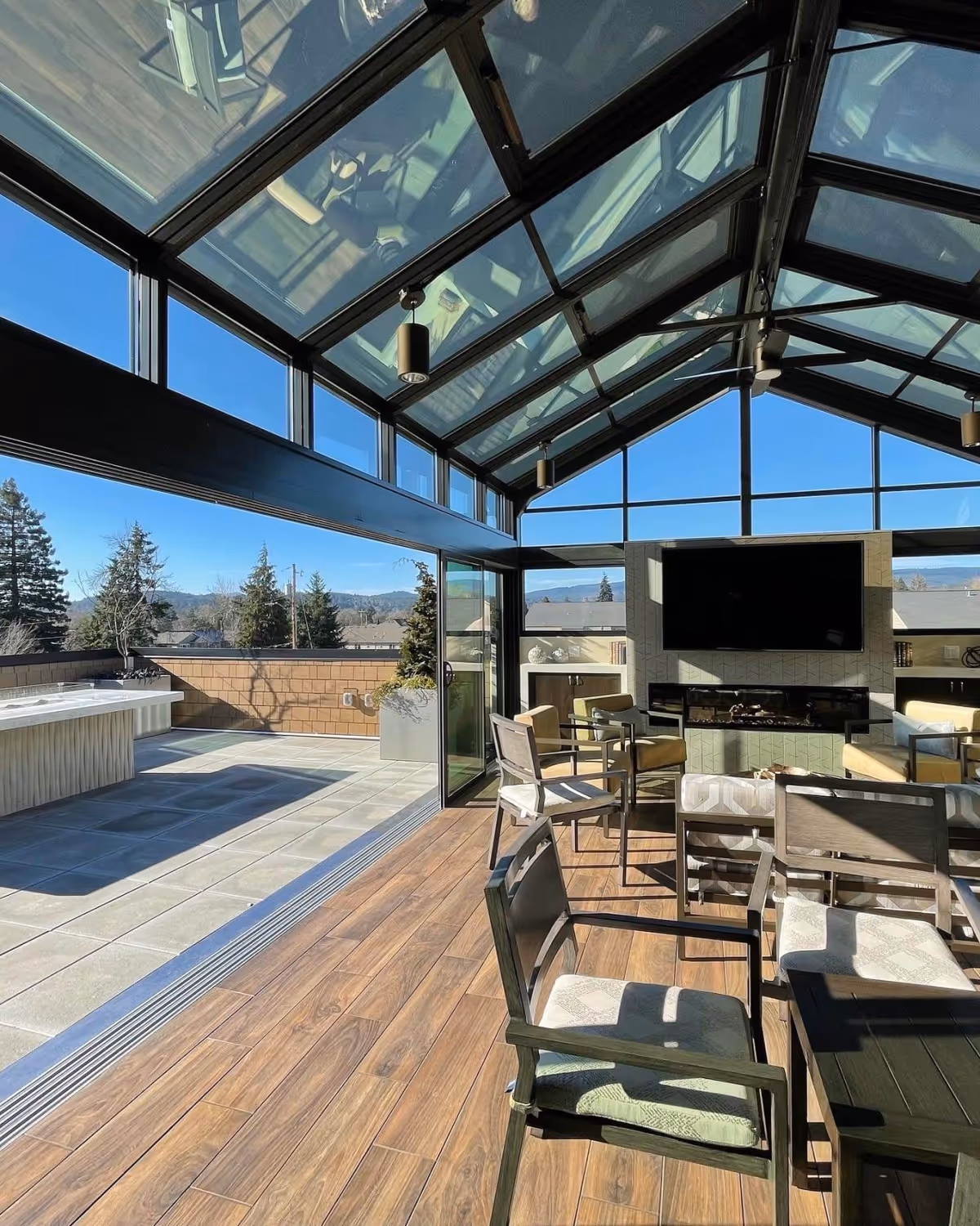Sunlit indoor-outdoor lounge with a glass roof, chairs and a TV opening onto a rooftop terrace with a hot tub.