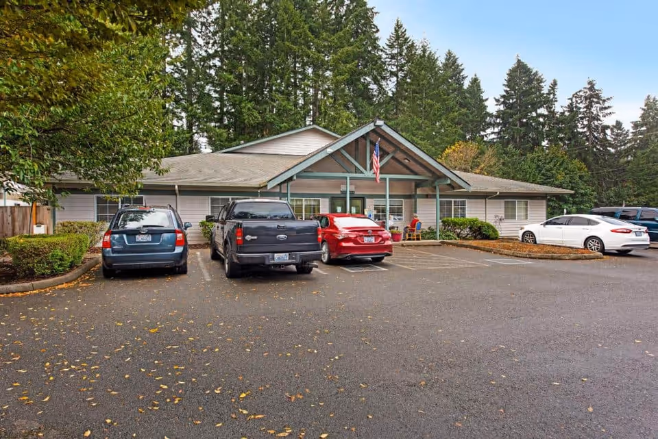Single-story memory care building with a covered entrance, parking spaces and several cars parked in front against a backdrop of tall trees.