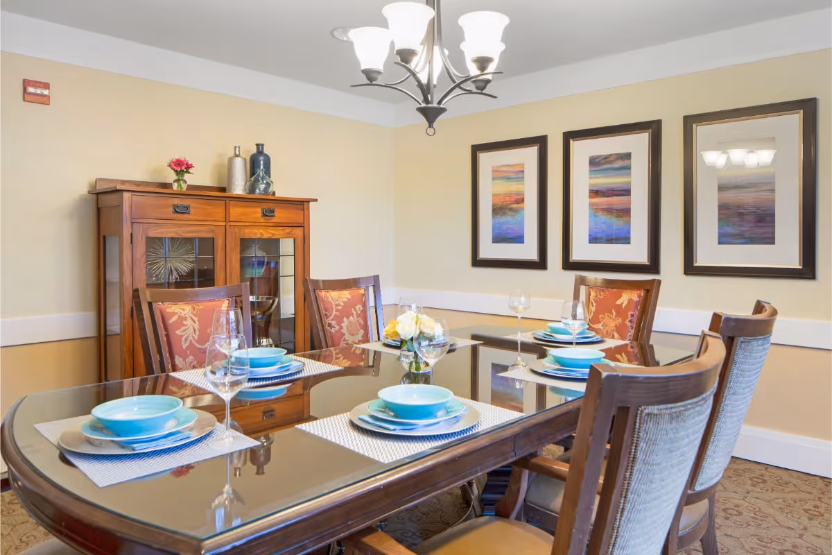 A dining room with a glass-topped table set with blue dishes, wooden chairs, a sideboard and framed artwork on the wall.