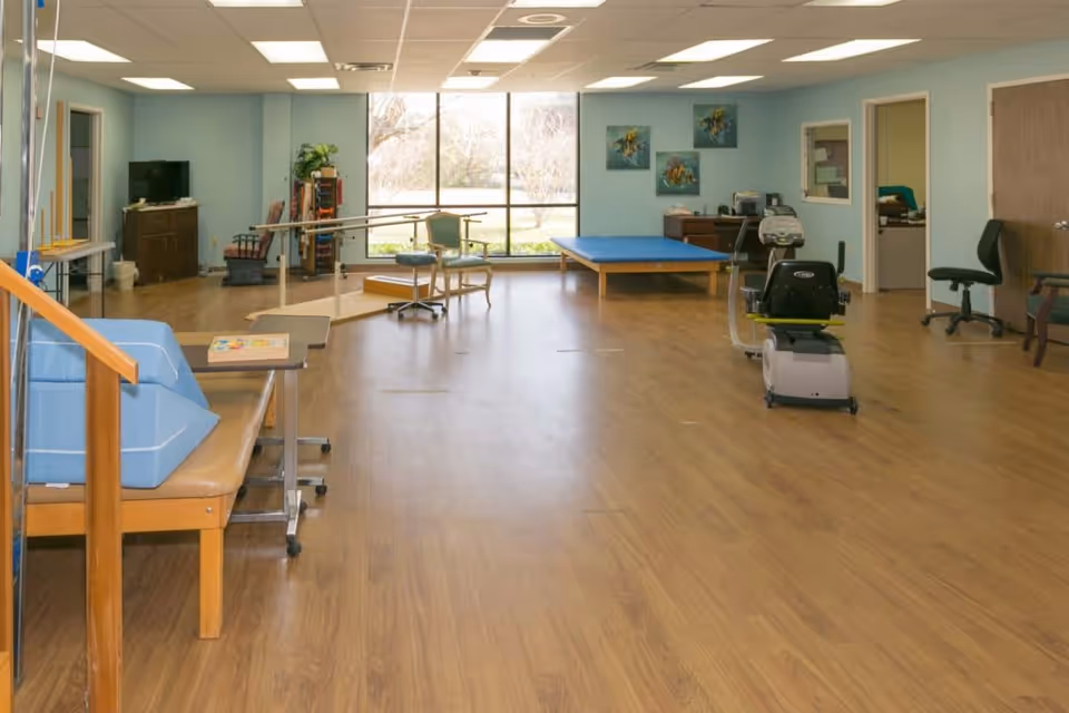A spacious room with wooden flooring and light blue walls, equipped with physical therapy and rehabilitation equipment including parallel bars, exercise bike, therapy tables, chairs, and a TV. Large windows provide natural light and a view of the outdoors.