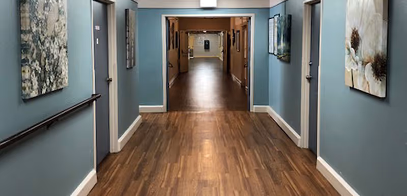 A long indoor hallway with wooden flooring and blue walls, featuring handrails on both sides and several closed doors. Floral paintings hang on the walls, and the hallway extends into another corridor in the distance.