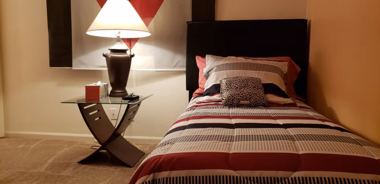 A neatly made single bed with a striped comforter in shades of red, white, black, and gray, and a leopard print pillow on top. Next to the bed is a modern glass-top nightstand with a dark brown base, holding a large table lamp with a white lampshade and a box of tissues. The room has beige walls and carpeted floor.