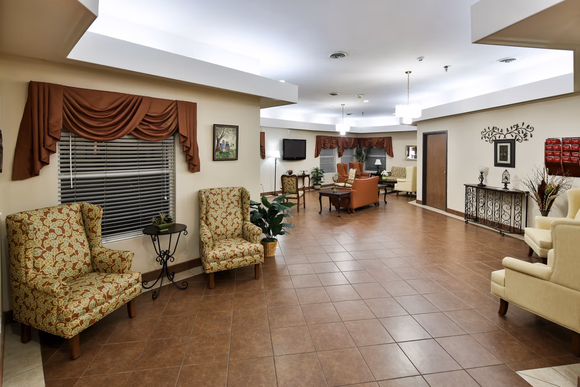 A spacious and well-lit common area in a nursing and rehabilitation center featuring multiple upholstered armchairs with patterned and solid fabrics, small tables, potted plants, wall decorations including framed pictures and a decorative metal wall art, and a wall-mounted television. The floor is tiled, and the windows have brown valances.