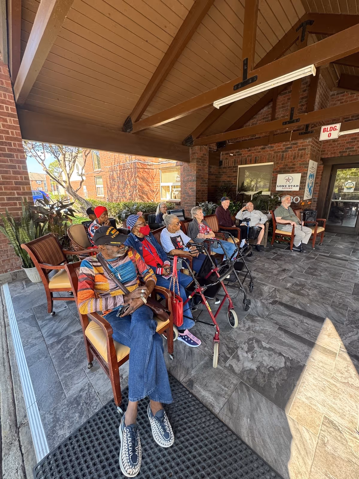 A group of elderly people sitting on chairs under a covered outdoor patio area at Lone Star Living. The patio has a wooden ceiling with exposed beams and a tiled floor. Some individuals have walkers and are dressed warmly. There are plants and a brick wall in the background with a Lone Star Living sign visible.