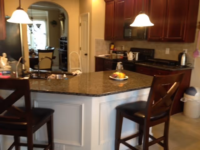 Interior view of a kitchen with dark wooden cabinets, a granite countertop island with two high chairs, pendant lights hanging above the island, and various kitchen appliances including a microwave, kettle, and toaster. In the background, an arched doorway leads to another room with a dining table and chairs.