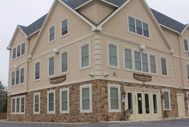 Exterior view of a three-story building with beige and stone facade, multiple windows, and a main entrance with double glass doors. Two signs on the building read 'Alexandria Manor'.