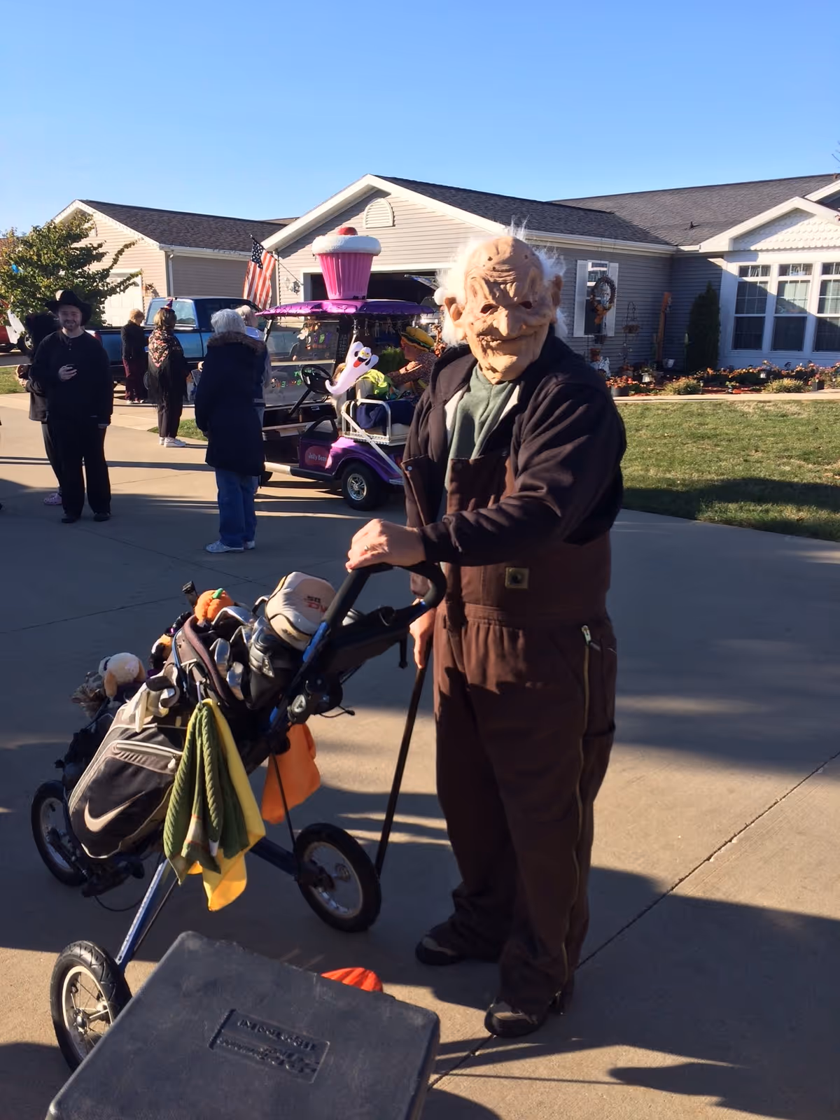 An elderly person wearing a creepy old man mask and brown overalls stands outside on a driveway, holding a golf push cart with golf clubs. In the background, there are several people, a decorated golf cart with a large pink cupcake on top, and a house with a well-maintained lawn under a clear blue sky.