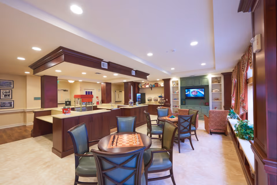 Bright communal dining and lounge area with tables and chairs, a serving counter, TV, and seating in a senior living facility.