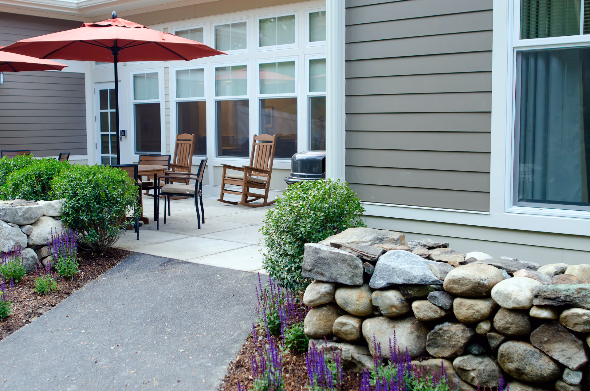 Outdoor patio with wooden rocking chairs, a table under a red umbrella, a stone wall and landscaping outside a senior living building.