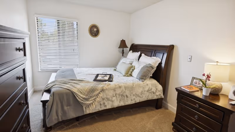 Neatly made bedroom with a wooden sleigh bed, matching dresser and nightstand, a lamp, and a window with blinds.