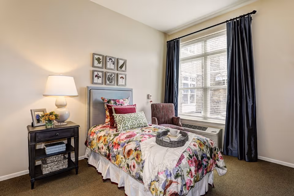 A cozy bedroom with a single bed covered in a colorful floral bedspread and multiple pillows. Next to the bed is a dark wooden nightstand with a white lamp, a small framed photo, and a vase of flowers. A comfortable armchair is placed near a large window with dark curtains, allowing natural light to fill the room.