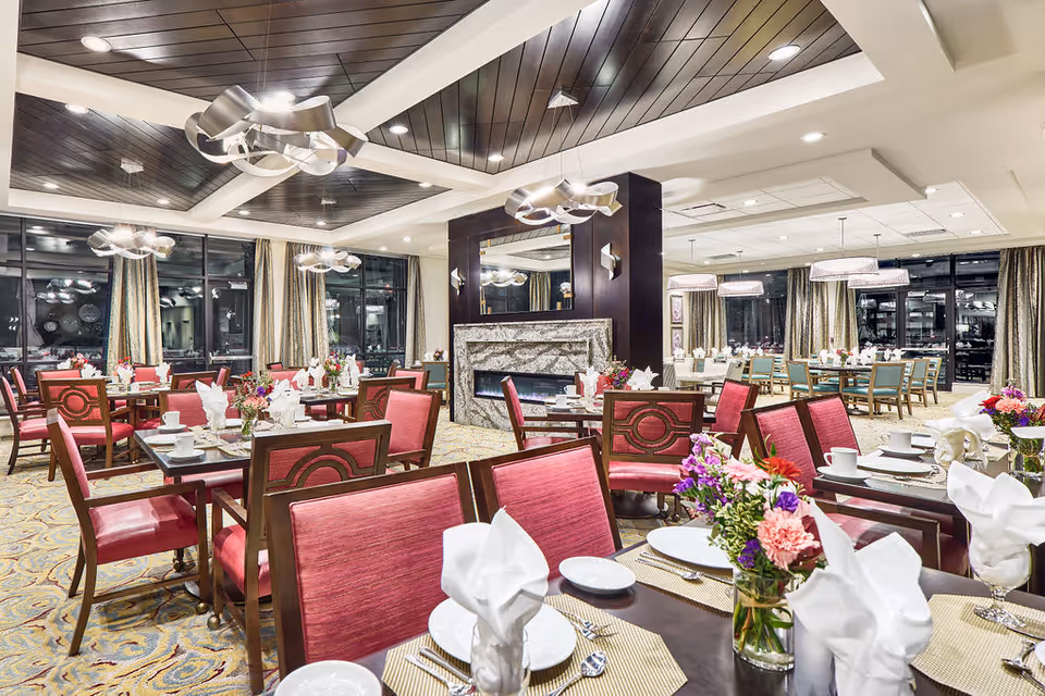 A spacious, well-lit dining room with multiple tables set with white plates, cups, silverware, and folded white napkins. The chairs have red upholstery, and there are flower arrangements on the tables. The room features large windows with curtains, modern ceiling lights, and a central fireplace with a dark wood panel above it.