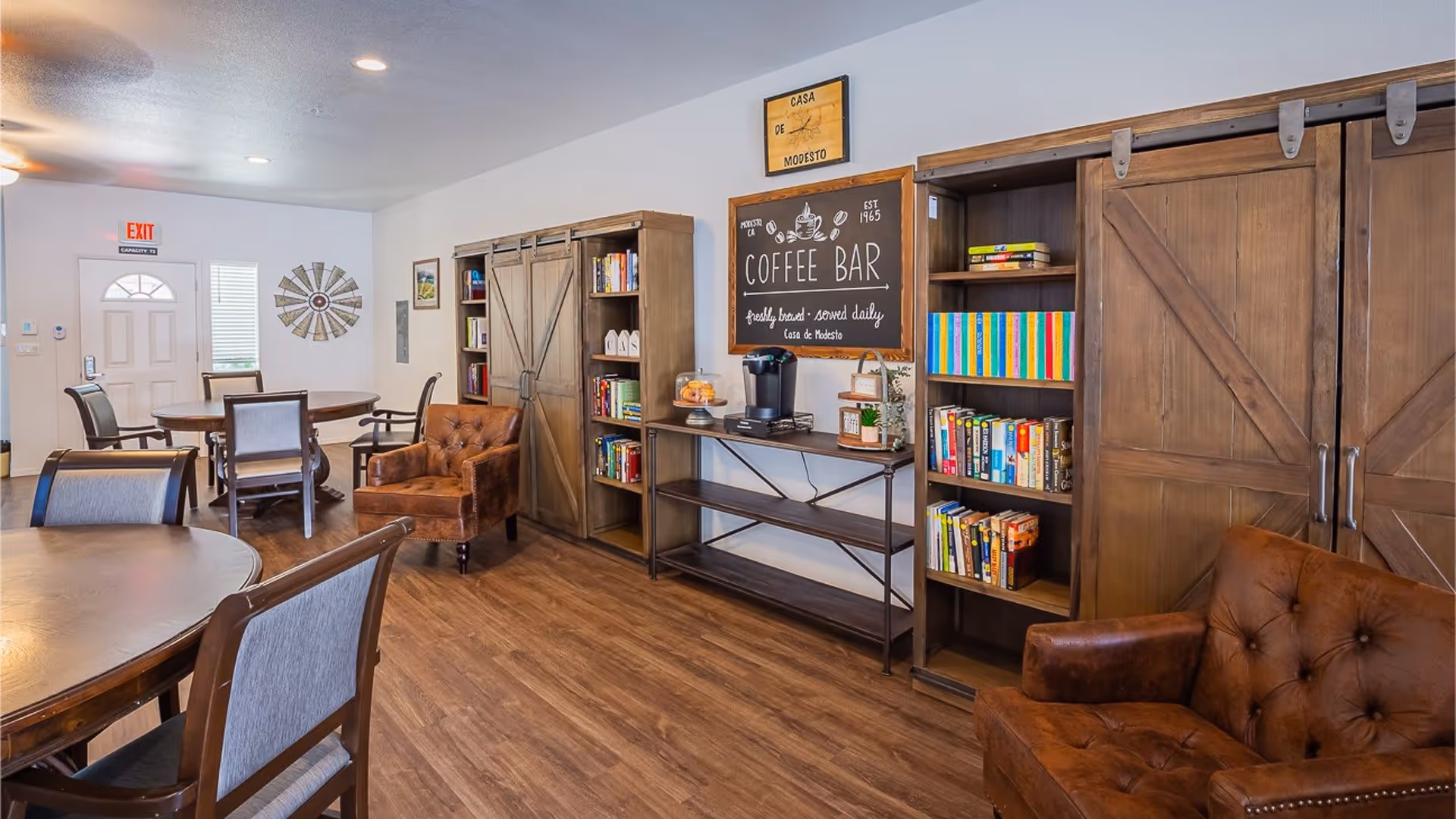 Interior view of a common area in Casa de Modesto featuring wooden tables and chairs, a brown leather armchair, bookshelves with books, a coffee bar with a coffee maker and pastries, and a decorative wall clock shaped like a windmill.