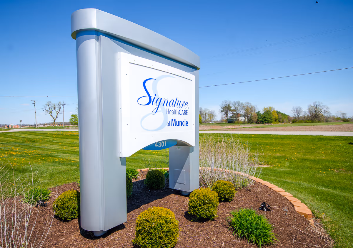 Outdoor view of a large silver and white sign for Signature HealthCARE of Muncie, surrounded by landscaped bushes and grass under a clear blue sky.