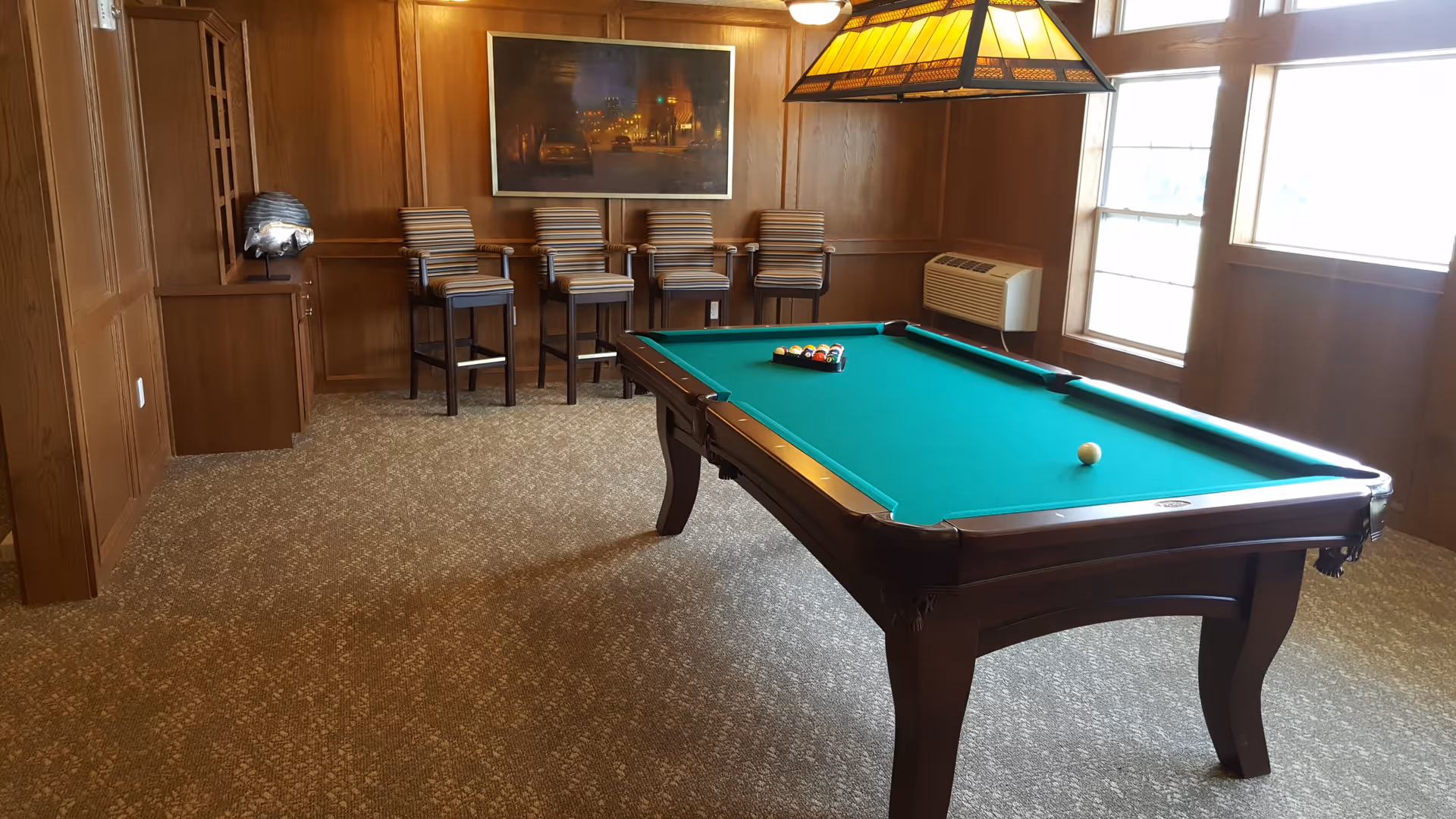 A room with wood-paneled walls featuring a pool table with a green felt surface and a rack of billiard balls set up for a game. There are four striped bar stools lined up against the back wall beneath a framed picture. A large window on the right side lets in natural light, and a hanging lamp with a stained glass shade is positioned above the pool table. The floor is carpeted in a neutral tone.
