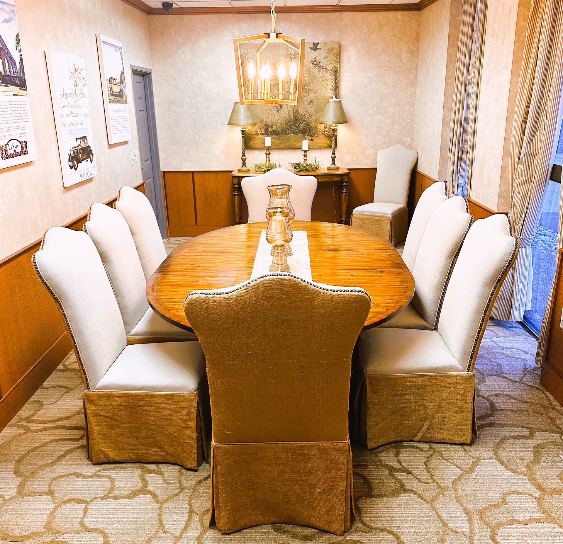A dining room with a wooden oval table surrounded by eight beige upholstered chairs. On the table are three glass candle holders. The room has beige walls with wood paneling on the lower half, a beige patterned carpet, and a window with striped curtains on the right. A side table with two lamps and decorative items is against the far wall, beneath a framed artwork. Three framed pictures hang on the left wall.